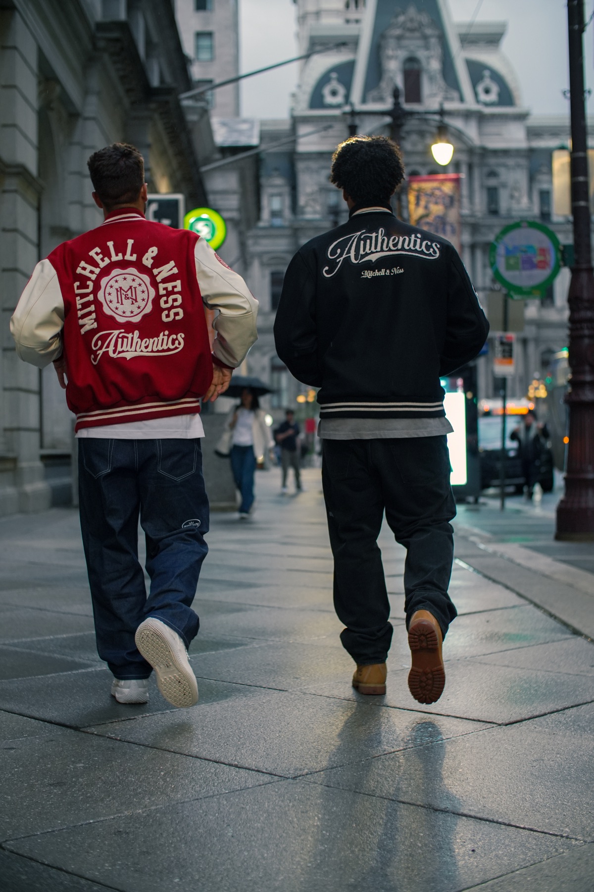 Two people in Mitchell & Ness varsity jackets walking a Philadelphia street toward City Hall